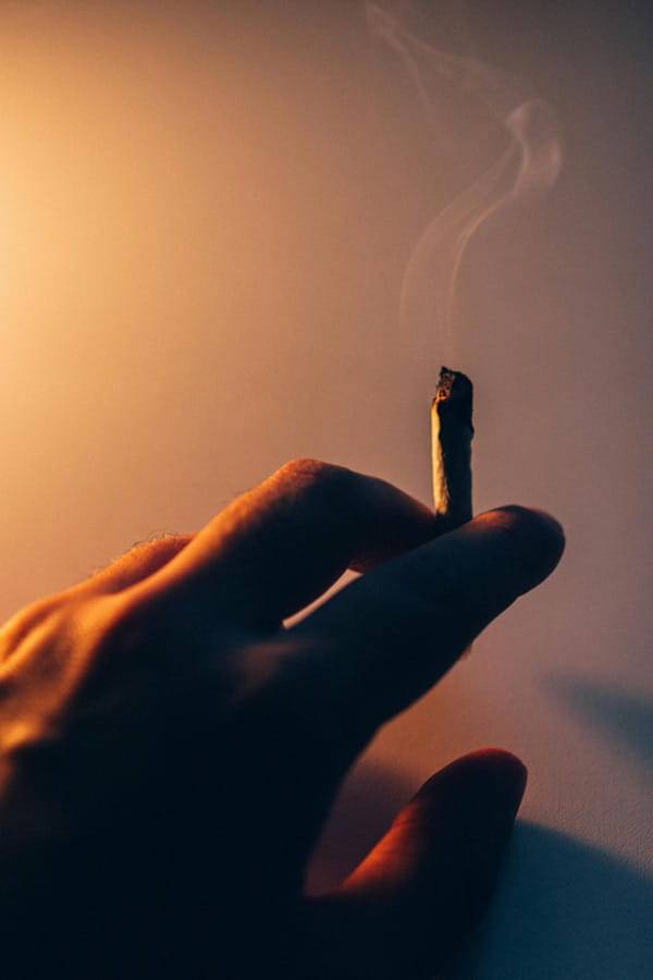 Hand hält einen rauchenden Joint vor warm beleuchtetem Hintergrund