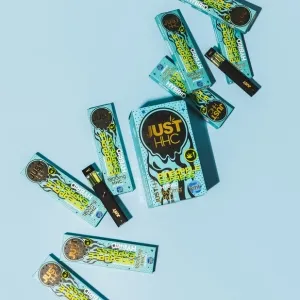 Mehrere hellblaue Produktverpackungen und schwarze Einweg-Vape-Pens der Sorte „Pineapple Express“ sind lose auf einem hellblauen Hintergrund verteilt. Die Verpackungen zeigen gelbe Schriftzüge und schwarze, tropfende Grafiken. Auf den Kartons steht „1800mg HHC“ und „Hybrid“.