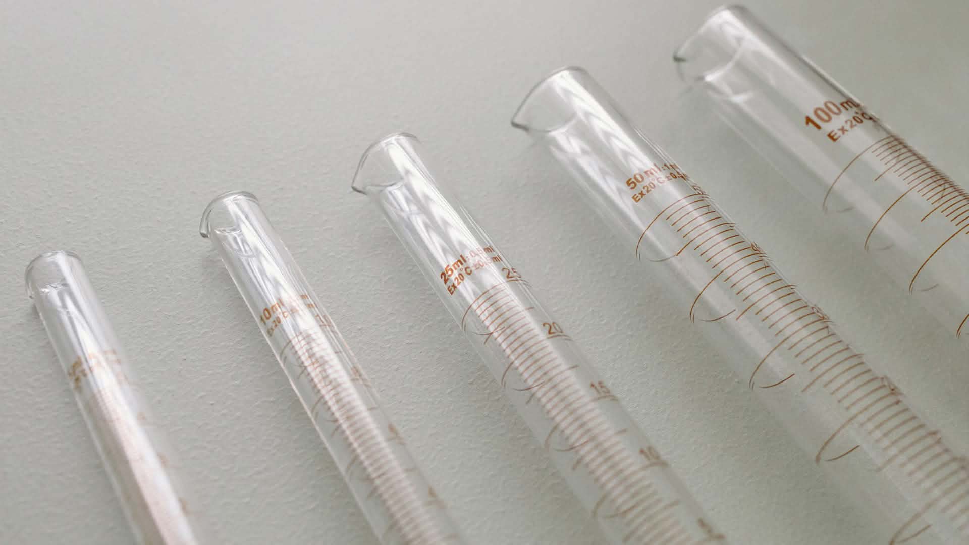 Mehrere unterschiedlich große Messzylinder aus Glas mit Volumenskalen liegen nebeneinander auf einer hellen Oberfläche